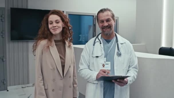 Retrato medio de médico maduro exitoso y paciente mujer feliz de pie en el vestíbulo del hospital moderno mirando a la cámara - Metraje, vídeo