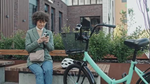 Mediana de largo tiro de la moderna mujer de negocios caucásica sentado en el banco con su bicicleta cerca de buscar información en Internet en el teléfono inteligente - Metraje, vídeo