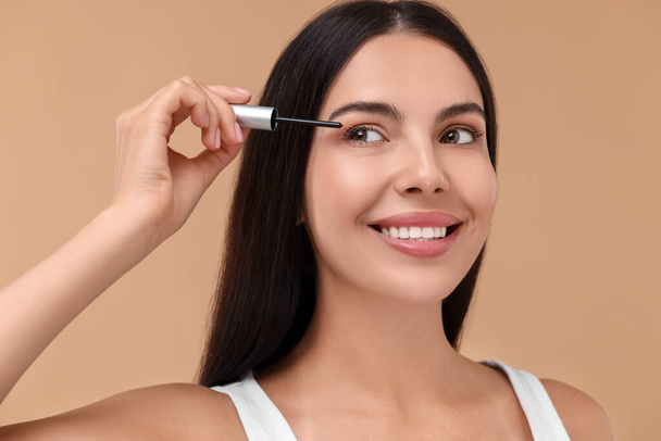 Beautiful woman applying serum onto her eyelashes on beige background. Cosmetic product - 写真・画像