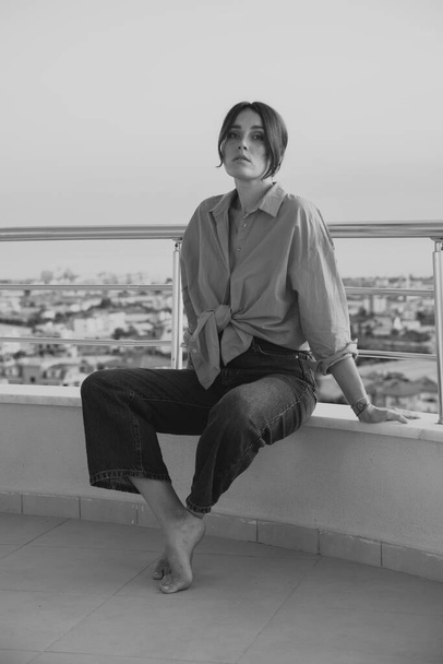 Hermosa chica en el balcón con vistas al mar. Una chica con camisa roja y vaqueros azules descansa en el balcón de su apartamento en un piso alto. - Foto, Imagen