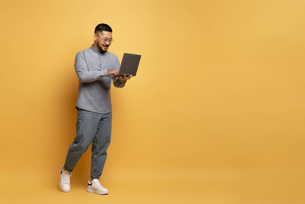 Vidám fiatal ázsiai férfi laptop séta sárga stúdió háttér, teljes hosszúságú lövés mosolygós Millennial Guy segítségével PC Számítógép távoli munka vagy online vásárlás, Másolás tér - Fotó, kép