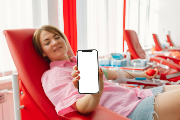 Donación de sangre. Primer plano de la mano femenina sosteniendo teléfono inteligente con pantalla en blanco en el centro de donación de sangre. Copiar espacio - Foto, Imagen