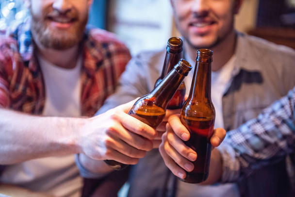 Rajattu kuva komeita ystäviä clinking pulloa olutta ja hymyillen kun lepää pubissa
 - Valokuva, kuva
