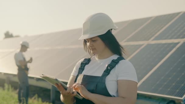 Egy női mérnök és asszisztense kemény kalapban egy megújuló energiaforrású erőműben, kezében egy táblagéppel és ellenőrzi a napelemek telepítését.Alternatív energia.Clean energy concept.4k - Felvétel, videó