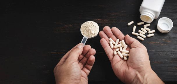 Male hands holds a scoop of whey or soy protein powder and white capsules of amino acids, vitamins, creatine, bodybuilding food supplements, sports nutrition on a dark wooden board. - Photo, Image
