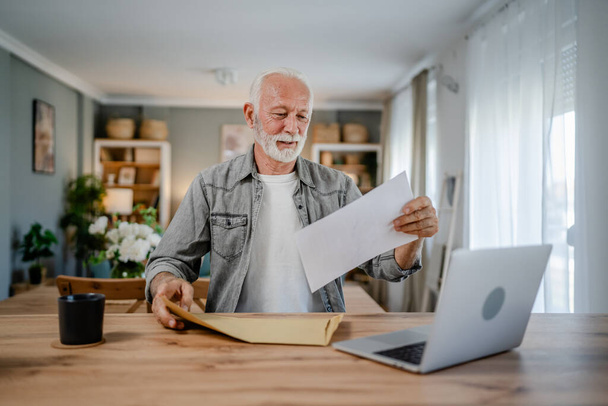 starszy kaukaski mężczyzna list otwarty poczta lub koperta dokumentu podczas siedzieć przy stole w domu otrzymując wyciąg bankowy kredyt zatwierdzenia umowy lub zaproszenie aktualności ubezpieczenia biznesu - Zdjęcie, obraz