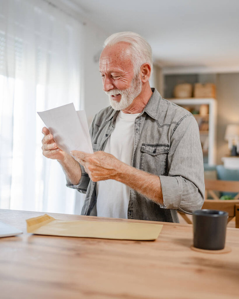 старший кавказький чоловік відкриває поштовий лист або документ конверт, сидячи за столом вдома отримуючи банківську виписку кредитний договір або запрошення на бізнес-страхування - Фото, зображення