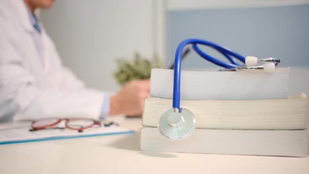 Vue rapprochée du stéthoscope avec livres sur table et médecin en utilisant une tablette sur fond d'hôpital - Séquence, vidéo