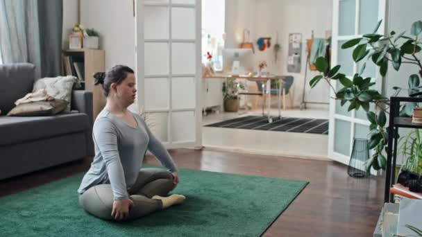Nowoczesna młoda kobieta z zespołem Downa siedzi na podłodze w salonie ćwicząc jogę - Materiał filmowy, wideo
