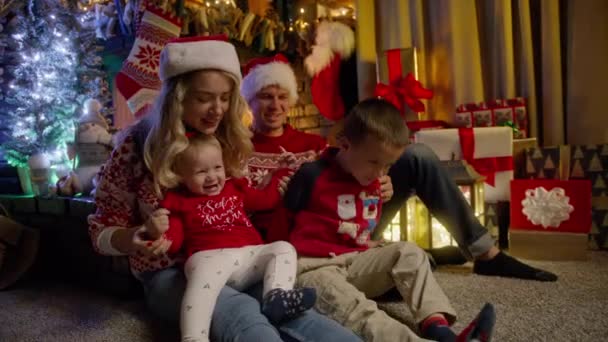 Щаслива сім'я зібралася напередодні Різдва біля ялинки. Молоді красиві батьки в червоних капелюхах Санти і маленька донька, дошкільнята, насолоджуються разом в затишному каміні RED 4K - Кадри, відео
