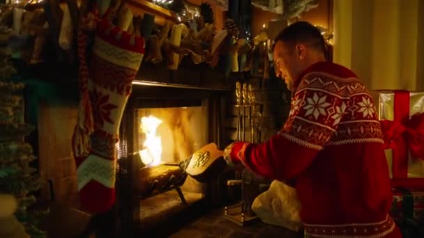 Young handsome man in Christmas sweater by cozy fireplace in mountain cabin. Smiling guy using bellows to blow air into fire. Winter season and Christmas holidays concept, slow motion RED camera shot - Footage, Video