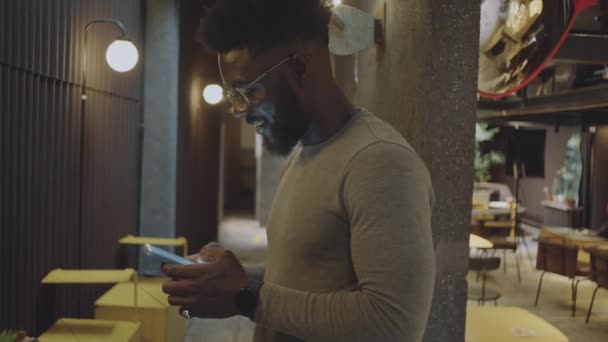 Μέτριο πλάνο του νεαρού μαύρου άνδρα σε casualwear και γυαλιά ηλίου στέκεται δίπλα καφέ τοίχο με σύγχρονο εσωτερικό και γραπτών μηνυμάτων σε smartphone - Πλάνα, βίντεο