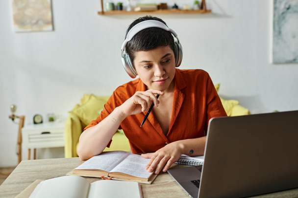 femme chère en chemise orange avec écouteurs et livre étudiant à son ordinateur portable à la maison, éducation - Photo, image