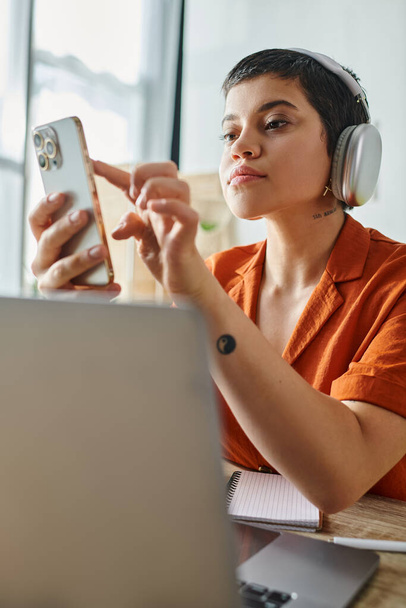 pionowe ujęcie atrakcyjnej studentki z krótkimi włosami ze słuchawkami patrzącymi na telefon przed laptopem - Zdjęcie, obraz