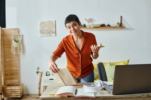 alegre estudiante atractiva en traje casual de pie junto al escritorio con su computadora portátil y libros de texto - Foto, Imagen