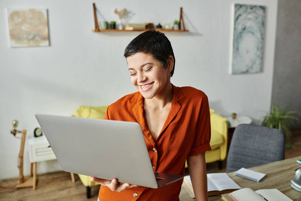 vrolijk jong vrouw met piercing in oranje shirt kijken blij op haar laptop, onderwijs - Foto, afbeelding