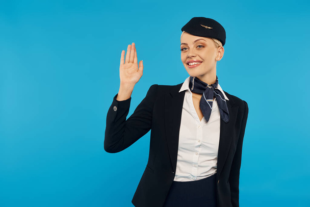 jeune hôtesse accueillante en uniforme professionnel élégant agitant la main sur fond de studio bleu - Photo, image
