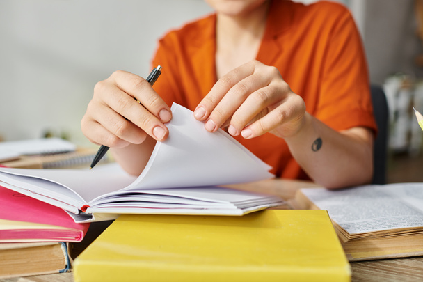 abgeschnittene Ansicht eines jungen Studenten mit Tätowierung in orangefarbenem Hemd, der Lehrbuchseiten blättert und von zu Hause aus lernt - Foto, Bild