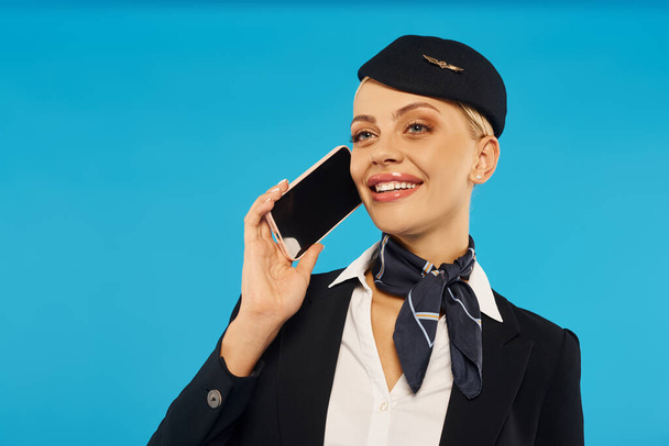 joven y alegre azafata en elegante uniforme hablando en smartphone con pantalla en blanco en azul - Foto, imagen