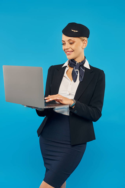 wesoła młoda kobieta w eleganckim mundurze stewardesy za pomocą laptopa, stojąc na niebieskim tle - Zdjęcie, obraz