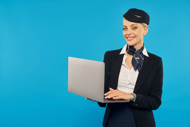 fröhliche Stewardess in eleganter Firmenuniform hält Laptop in der Hand und blickt in die Kamera auf blauem Hintergrund - Foto, Bild