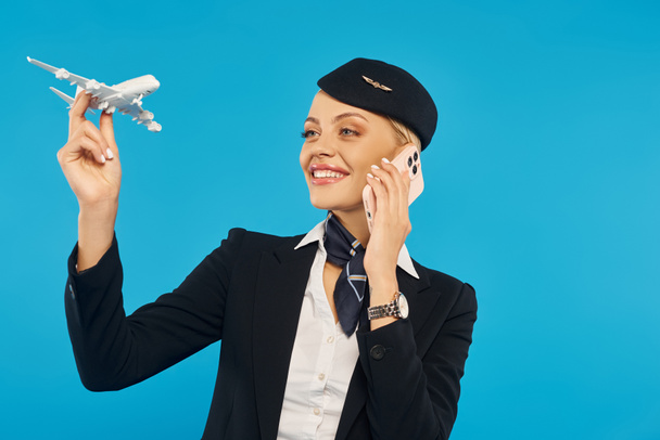 щаслива стюардеса в уніформі тримає модель літака і говорить om смартфон на синьому фоні - Фото, зображення