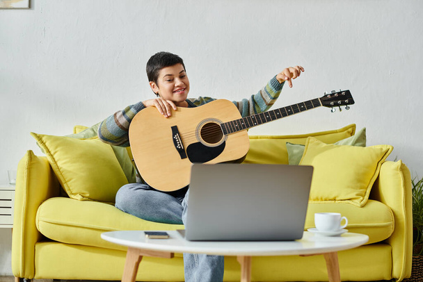 νεαρή χαρούμενη γυναίκα σε casual ενδυμασία κάθεται στον καναπέ μαθαίνοντας πώς να παίξει κιθάρα σε απομακρυσμένη τάξη - Φωτογραφία, εικόνα
