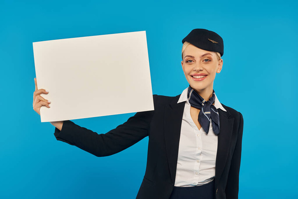 giovane hostess allegra in elegante uniforme in piedi con poster vuoto su sfondo blu studio - Foto, immagini