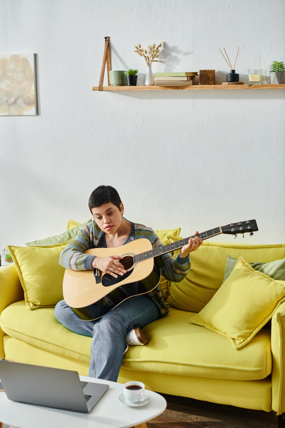 vertikale Aufnahme einer fokussierten jungen Frau, die mit Gitarre auf dem Sofa sitzt und an einer Online-Musikstunde teilnimmt - Foto, Bild
