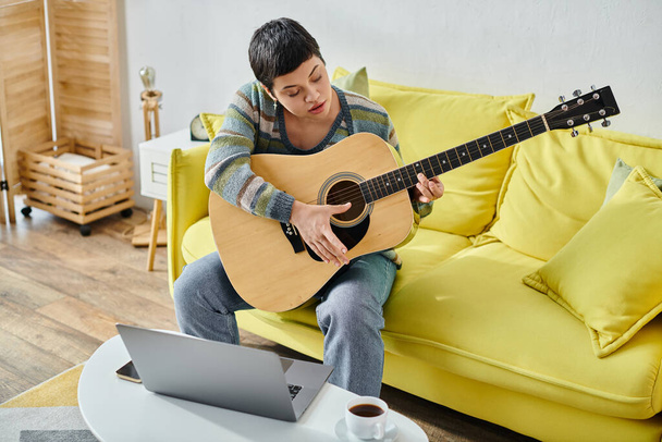 сконцентрированная молодая женщина, сидящая на диване, практикуя гитару на дистанционном уроке музыки, образования - Фото, изображение