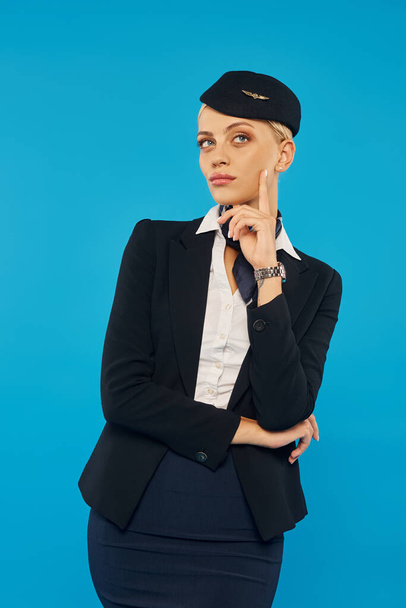 giovane e sognante donna in uniforme di hostess aerea toccare il viso e distogliere lo sguardo sullo sfondo blu - Foto, immagini