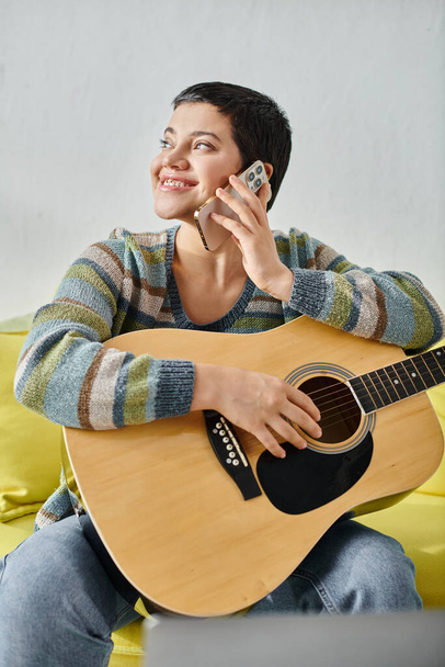 κάθετη λήψη χαρούμενη γυναίκα με σκουλαρίκια κρατώντας κιθάρα και μιλώντας από το τηλέφωνο, εκπαίδευση στο σπίτι - Φωτογραφία, εικόνα