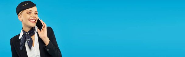 iloinen lentoemäntä tyylikkäässä univormussa puhuu matkapuhelimella sinisellä taustalla, vaakasuora banneri - Valokuva, kuva