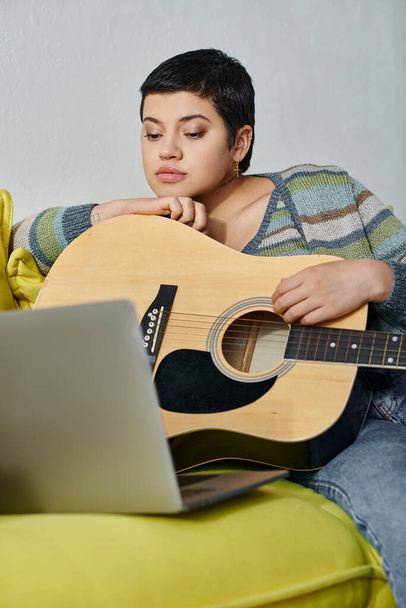 vertikale Aufnahme einer jungen attraktiven Frau, die auf dem Sofa sitzt und an einer Online-Gitarrenstunde teilnimmt - Foto, Bild