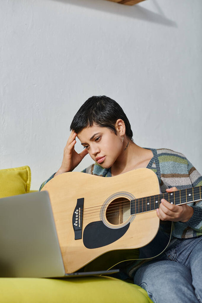 εστιασμένη κουρασμένη γυναίκα σε casual ενδυμασία μαθαίνοντας πώς να παίξει κιθάρα και κοιτάζοντας φορητό υπολογιστή, εκπαίδευση - Φωτογραφία, εικόνα