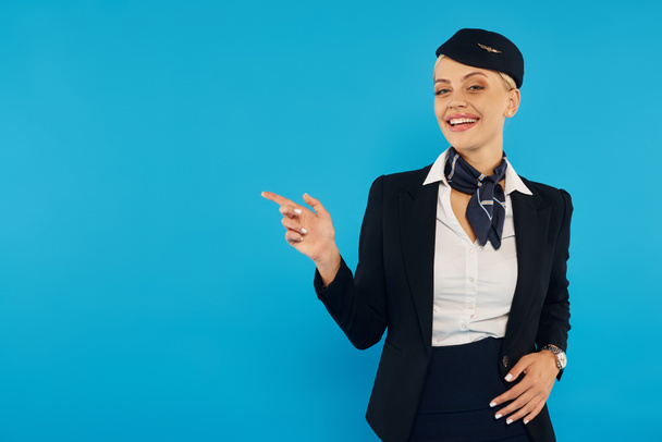 Lächelnde Stewardess zeigt weg, während sie die Richtung angibt und in die Kamera auf blauem Hintergrund schaut - Foto, Bild