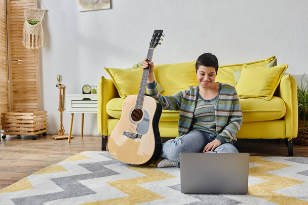 good looking woman in casual homewear sitting on floor with guitar and laptop at remote music class - Photo, Image