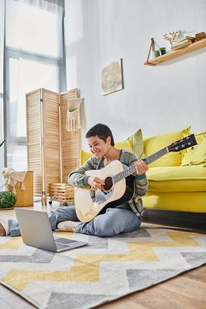 plan vertical de la femme assise sur le sol avec la guitare souriant à la caméra de l'ordinateur portable, éducation à la maison - Photo, image
