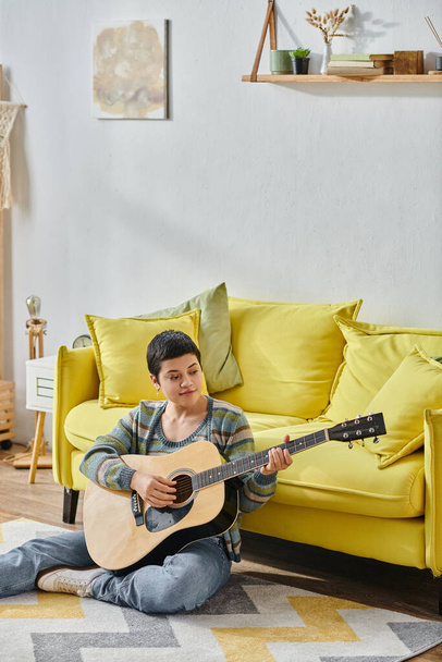 κάθετη βολή της εμφανίσιμης νεαρής γυναίκας που παίζει κιθάρα και κάθεται στο πάτωμα, εκπαίδευση στο σπίτι - Φωτογραφία, εικόνα