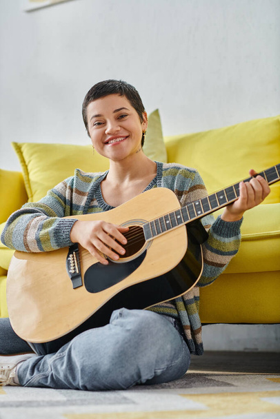 colpo verticale di donna allegra seduta sul pavimento con la chitarra sorridente alla macchina fotografica, educazione a casa - Foto, immagini