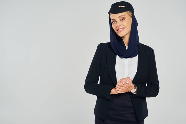 radosna stewardessa w chustce i elegancki mundur arabskich linii lotniczych uśmiechających się do kamery na szarości - Zdjęcie, obraz