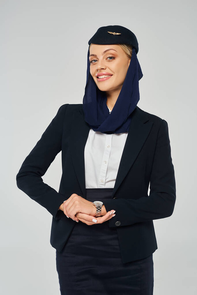 sierlijke stewardess van Arabische luchtvaartmaatschappijen met een vrolijke glimlach en hoofddoek kijkend naar camera op grijs - Foto, afbeelding
