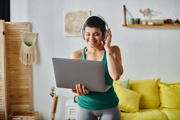 cheerful short haired woman with headphones looking at laptop camera during online fitness lesson - Photo, Image