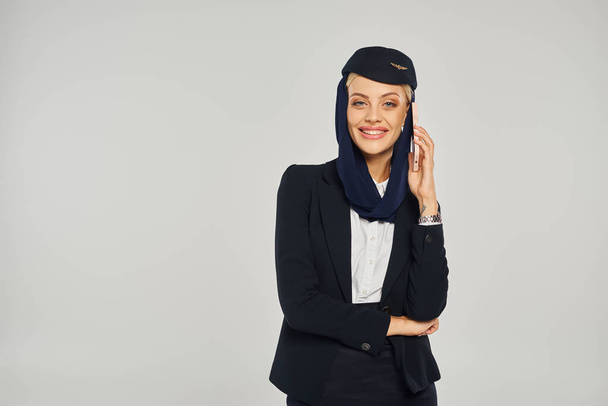 gracieuse hôtesse de l'air des compagnies arabes parlant sur téléphone portable et souriant à la caméra sur gris - Photo, image
