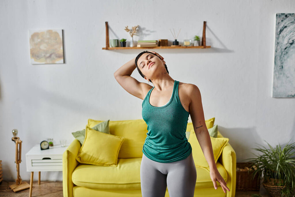 dobrze wyglądająca młoda kobieta z krótkimi włosami rozciągająca swoje ciało przed żółtą sofą, fitness - Zdjęcie, obraz