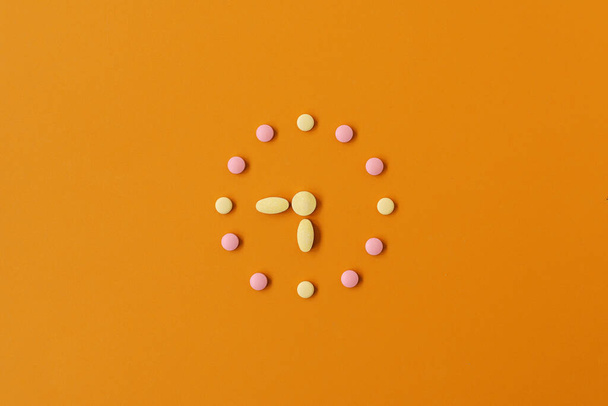 Ρολόι δημιουργήθηκε από ροζ και κίτρινο χάπια σε παστέλ πορτοκαλί φόντο. Η κατάλληλη στιγμή για τη χρήση φαρμάκων. Έννοια της υγειονομικής περίθαλψης και θεραπείας - Φωτογραφία, εικόνα