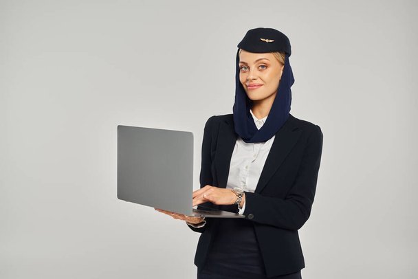 gelukkig Arabisch luchtvaartmaatschappijen stewardess in elegante uniform holding laptop terwijl staan op grijs - Foto, afbeelding
