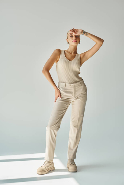stijlvolle blonde vrouw in tank top en broek poseren met de hand in de buurt voorhoofd op grijs, fashion shoot - Foto, afbeelding