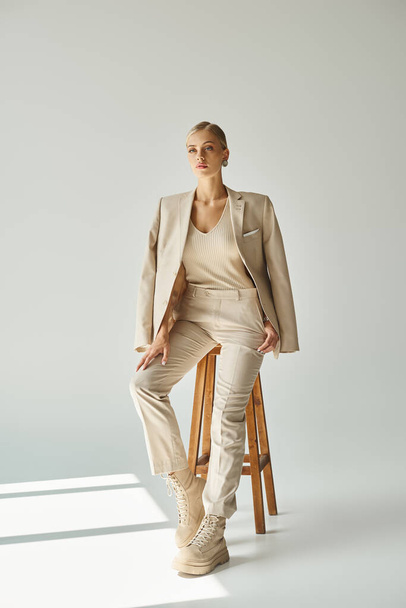 dreamy young woman in pastel beige suit looking away on tall stool in sunlight on grey backdrop - Фото, изображение