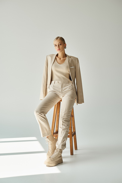 pretty blonde woman in pastel beige suit looking at camera on tall stool in sunshine on grey - Photo, Image